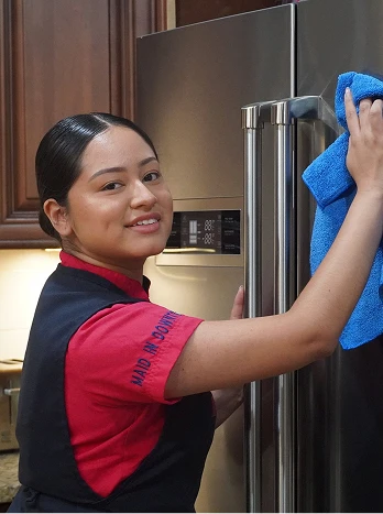 Woman in red shirt cleaning stainless steel refrigerator in modern kitchen with wooden cabinets - Cleaning in the DC Metro area.