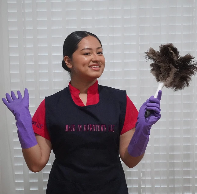 Professional housekeeper in uniform holding duster and wearing purple gloves, smiling at camera - in the DC Metro area.