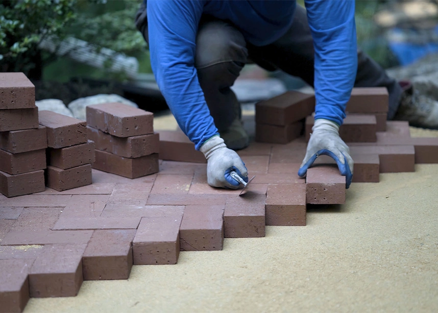 Worker installing red brick pavers in herringbone pattern on sand base for patio or walkway construction - Hardscaping in the DC Metro area.