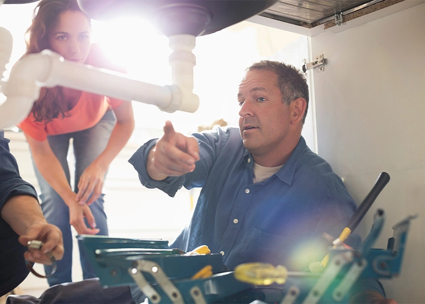 Man showing plumbing to woman under the sink. He is wearing a blue work shirt and is pointing at a white PVC pipe. - Plumbing in the DC Metro area.