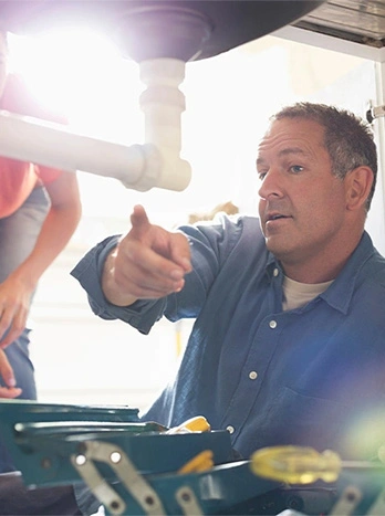 Man showing plumbing to woman under the sink. He is wearing a blue work shirt and is pointing at a white PVC pipe. - Plumbing in the DC Metro area.